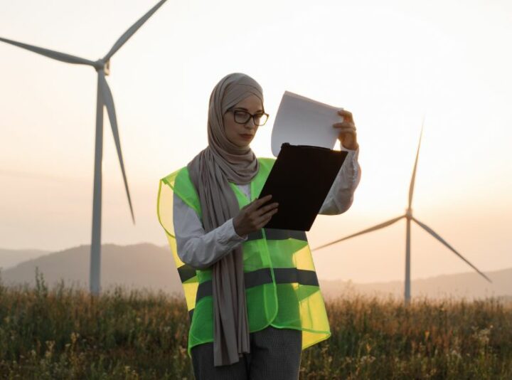 A lady in a high-vis jacket and head scarf looking at a clipboard in front of 2 wind turbines