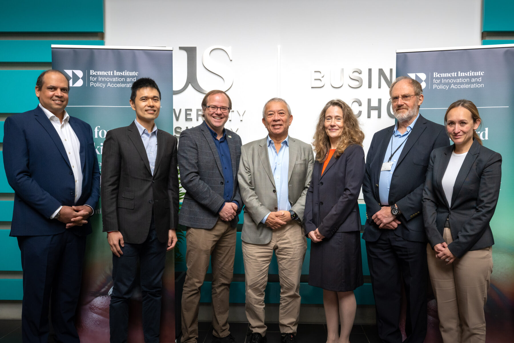 Seven people smiling at the camera in front of Business School sign and Bennett Institute pop-up banners